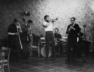 A jazz band Left to right: Man playing trombone, man playing double bass, man playing trumpet, man playing drum kit, man playing clarinet, man playing banjo